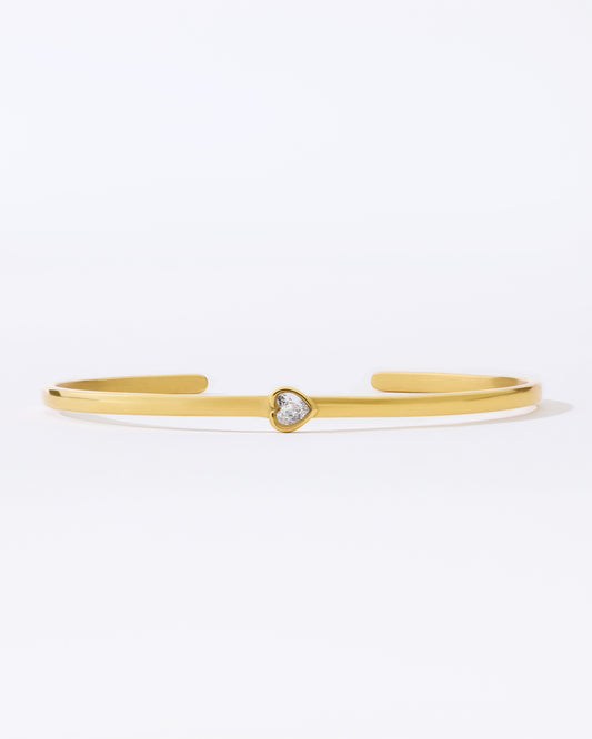 Gold bangle bracelet with a small heart shaped gemstone on a white background